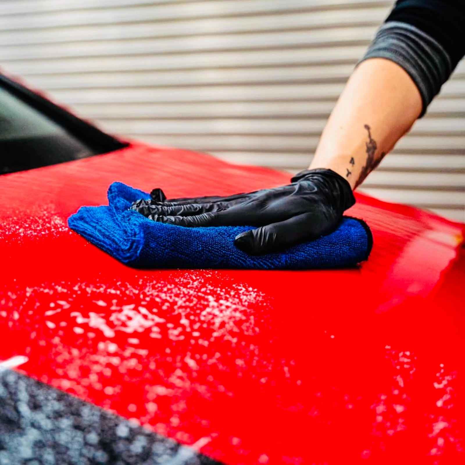 Gloved hand using a blue microfiber towel to wipe a red vehicle surface during automotive detailing.